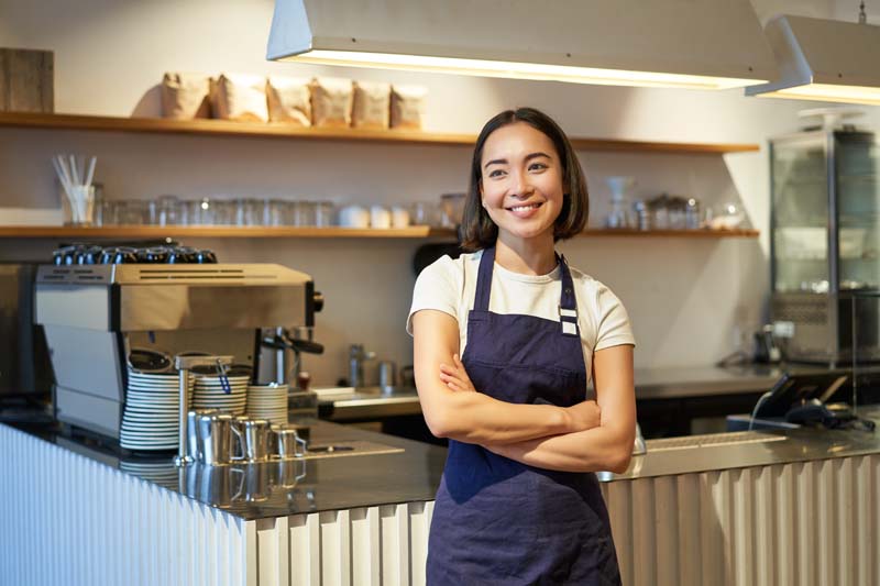 Smiling asian girl barista, standing near counter in cafe, wearing company apron, cross hands and looks confident